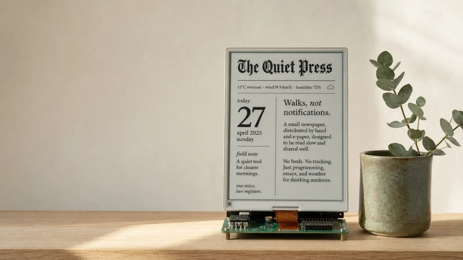 The e-paper press on the shelf in soft morning light, its screen showing the masthead over a single line, 'Walks, not notifications', and a short note: a small newspaper, distributed by hand and e-paper, designed to be read slow and shared well. A sprig of eucalyptus in a stoneware cup stands beside it.