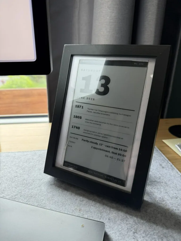 The weekend edition running for real: a framed e-paper panel standing on a desk in daylight, showing a heavy sans-serif Saturday 13, three dated history briefs, a weather line reading partly cloudy, 12 degrees, rain from 14:00, a line noting one appointment at 20:00, and sun times 04:40 to 21:27. A monitor and curtained window sit softly out of focus behind it.